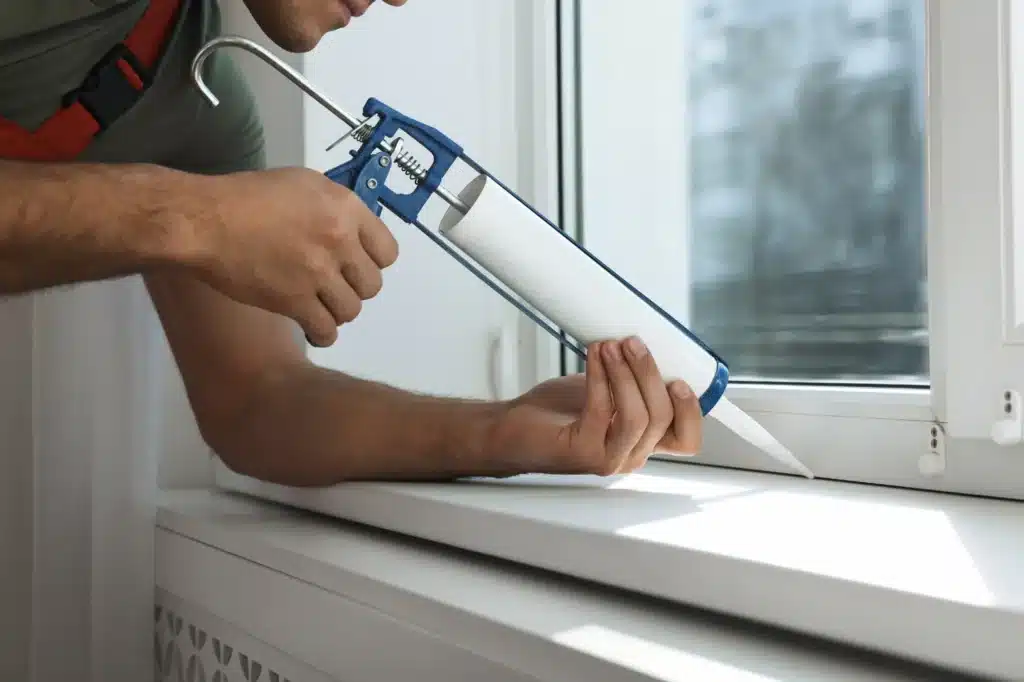 person using a caulking gun to apply sealant around the edge of a window frame 