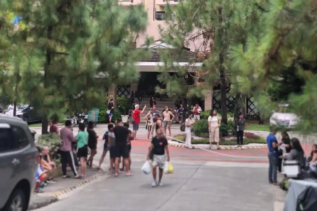 People gathered outside a building, possibly during an evacuation or emergency drill, surrounded by trees and parked vehicles.