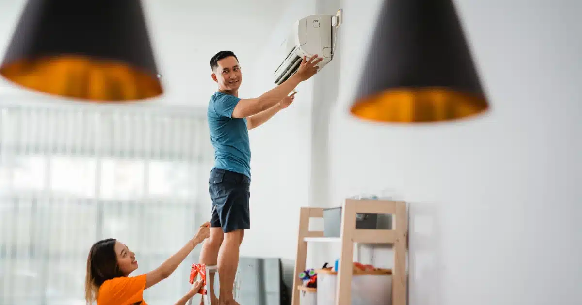 A man standing on a ladder handling a wall-mounted air conditioner while a woman below steadies the ladder inside a bright, modern room.