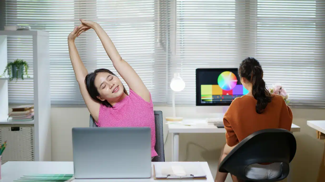An office worker stretching her arms overhead at her desk while sitting in front of a laptop, with a coworker and a monitor displaying colorful graphics in the background.